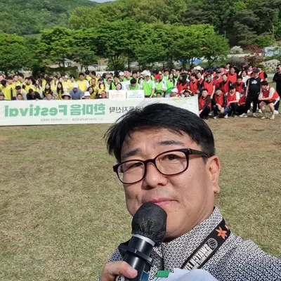 야외 행사장에서 마이크를 들고 참가자들을 배경으로 한 신용주 MC