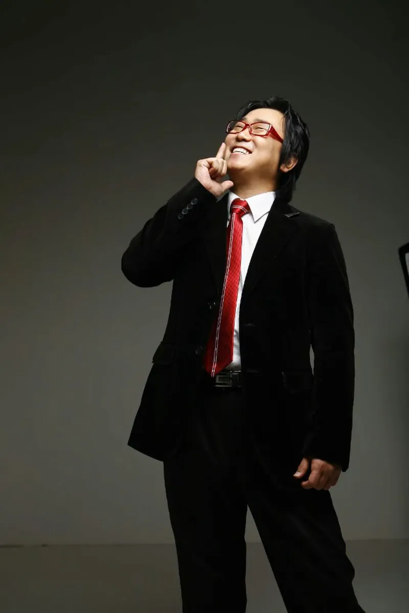 검은 정장과 붉은 넥타이 차림으로 서 있는 신용주 MC의 스튜디오 컷